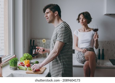 Young Sad Couple Two Woman Man 20s Wearing Casual T-shirt Clothes Prepare Vegetable Salad Girl Sit On Table Drink Red Wine Cook Food In Light Kitchen At Home Together. Healthy Diet Lifestyle Concept.