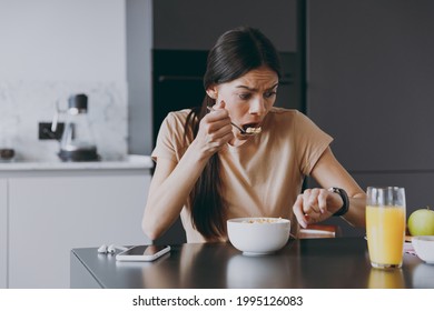 Young sad displeased disappointedhousewife woman 20s in t-shirt eat breakfast in morning look at smart watch check time hurry rush cooking food in light kitchen at home Healthy diet lifestyle concept