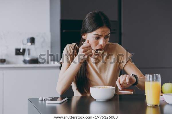 Young sad displeased disappointedhousewife woman\
20s in t-shirt eat breakfast in morning look at smart watch check\
time hurry rush cooking food in light kitchen at home Healthy diet\
lifestyle concept