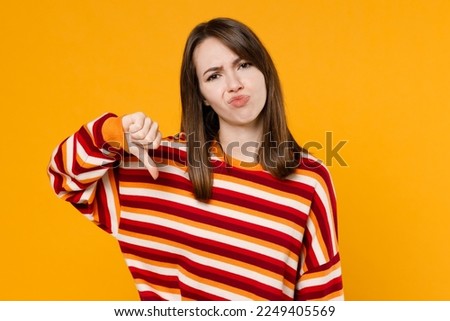 Young sad displeased dissatisfied caucasian woman 20s wearing red striped sweatshirt showing thumb down dislike gesture isolated on plain yellow background studio portrait. People lifestyle concept