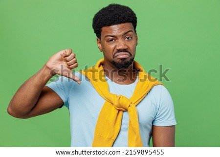 Young sad dissatisfied displeased man of African American ethnicity 20s in blue t-shirt showing thumb down dislike gesture isolated on plain green background studio portrait. People lifestyle concept