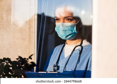 Young sad female caucasian UK EMS doctor carer looking through ICU window,fear and uncertainty in eyes,wearing face mask gazing at distance,hope in overcoming Coronavirus COVID-19 pandemic crisis