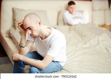 Young Sad Gay Man Sitting On Bed With His Lover Under Blanket On Background