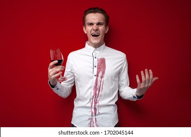 Young Sad Guy In A White Shirt Spilled Red Wine On Himself On A Red Background, An Angry Man Put A Stain Of Wine On His Clothes