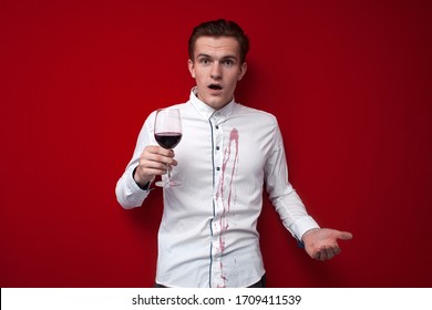 Young Sad Guy In A White Shirt Spilled Red Wine On Himself On A Red Background, An Angry Man Put A Stain Of Wine On His Clothes