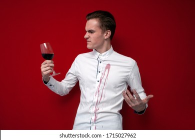 Young Sad Guy In A White Shirt Spilled Red Wine On Himself On A Red Background, An Angry Man Put A Stain Of Wine On His Clothes