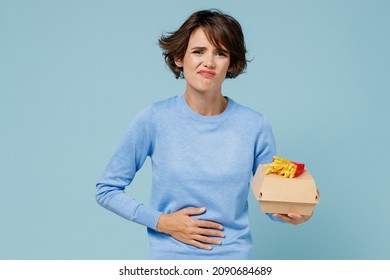 Young Sad Hungry Sick Woman Wearing Sweater Hold A Box With Fastfood French Fries Potato Burger Put Hand On Belly Isolated On Plain Pastel Light Blue Background. People Lifestyle Junk Food Concept.