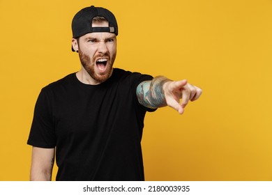 Young sad indignant angry bearded tattooed man 20s he wears casual black t-shirt cap point index finger aside scream isolated on plain yellow wall background studio portrait. People lifestyle concept