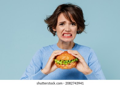 Young Sad Irritated Stressed Woman 20s Wearing Casual Sweater Look Camera Biting Eating Burger Isolated On Plain Pastel Light Blue Color Background Studio Portrait. People Lifestyle Junk Food Concept