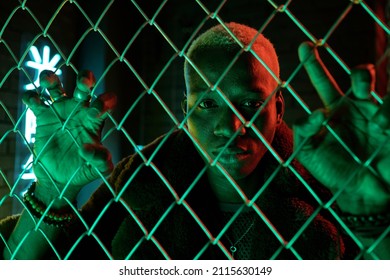 Young Sad Man Of African Ethnicity Standing Behind Bars And Looking At You Against Illuminated Hieroglyph In Darkness