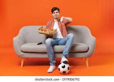 Young sad man football fan in shirt support team with soccer ball sit on sofa at home watch tv live stream eat pizza show thumb down gesture isolated on orange background People sport leisure concept - Powered by Shutterstock