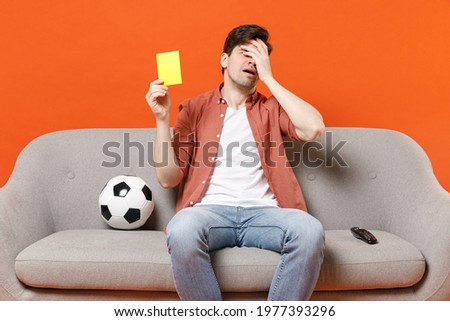 Young sad man football fan in shirt support team with soccer ball sit home sofa watch tv live stream put hand face facepalm gesture hold yellow card isolated on orange background People sport concept