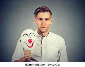 Young Sad Man With Happy Clown Mask Isolated On Gray Wall Background. Human Emotions