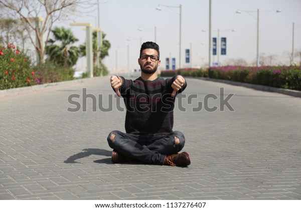 Young\
sad man sitting crossed legs on the street thumbs down with\
frowning face, on the road of the cars in the\
mall.