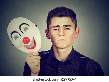 Young Sad Man Taking Off Happy Clown Mask Isolated On Gray Wall Background. Human Emotions. Split Personality Concept