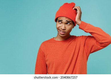 Young sad puzzled confused embarrassed african american man 20s in orange shirt hat look aside scratch head isolated on plain pastel light blue background studio portrait. People lifestyle concept.
