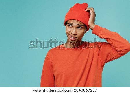 Young sad puzzled confused embarrassed african american man 20s in orange shirt hat look aside scratch head isolated on plain pastel light blue background studio portrait. People lifestyle concept.