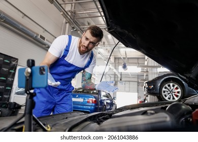 Young Sad Puzzled Technician Car Mechanic Man In Blue Overalls White T-shirt Talk Mobile Cell On Phone Stand Asking How To Do Fix Problem With Raised Hood Work In Vehicle Repair Shop Workshop Indoors.