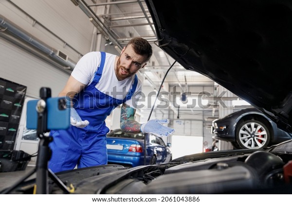 Young sad puzzled technician car mechanic man in\
blue overalls white t-shirt talk mobile cell on phone stand asking\
how to do fix problem with raised hood work in vehicle repair shop\
workshop indoors.