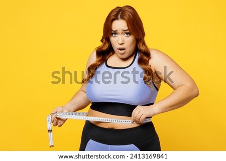 Young sad shocked chubby overweight plus size big fat fit woman wearing blue top warm up training hold measure tape on waist isolated on plain yellow background studio home gym. Workout sport concept