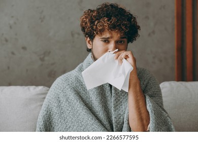 Young sad sick ill Indian man wapped in plaid sneezing have runny nose look camera sits on grey sofa couch stay at home hotel flat rest relax spend free spare time in living room indoor Lounge concept
