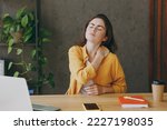 Young sad tired employee business woman 20s she wear casual yellow shirt put hand on shoulder neck close eyes suffer fro pain sit work at wooden office desk with pc laptop. Achievement career concept