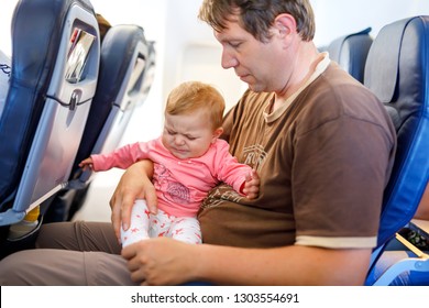 Young Sad Tired Father And His Crying Baby Daughter During Flight On Airplane Going On Vacations. Dad Holding Baby Girl On Arm. Air Travel With Baby, Child And Family Concept. Baby Wiht Earache.
