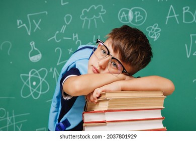 Young sad tired male kid school boy 5-6 years old in t-shirt backpack glasses lying on books isolated on green wall chalk blackboard background. Childhood children kids education lifestyle concept.