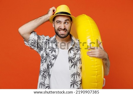Young sad tourist man wear beach shirt hat hold inflatable ring travel abroad on weekends hold scratch head isolated on plain orange background studio portrait Summer vacation sea rest sun tan concept