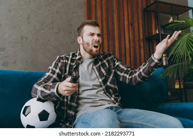 Young Sad Unhappy Man Fan In Brown Shirt Hold Remote Controller Spread Hands Cheer Up Support Football Team Sit On Blue Sofa With Soccer Ball Rest Watch Tv Indoors Room Gray Wall Sport Leisure Concept