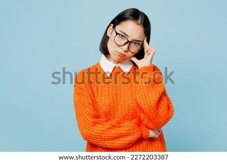 Young sad upset depressed woman of Asian ethnicity wearing orange sweater glasses look camera prop up head hold forehead isolated on plain pastel light blue cyan background. People lifestyle concept