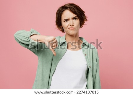Young sad woman 20s she wear green shirt white t-shirt look camera show thumb up dislike gesture reject refuse isolated on plain pastel light pink background studio portrait. People lifestyle concept.