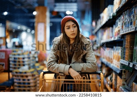 Young sad woman with empty shopping cart among produce aisle at supermarket.