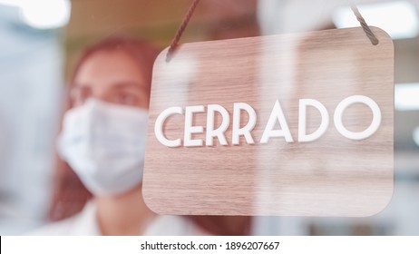 Young Sad Woman With Face Mask Changing Open To Closed Sign In Spanish On Window For Corona Virus