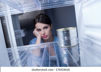 Young Sad Woman Looking At One Tin In Her Empty Fridge.