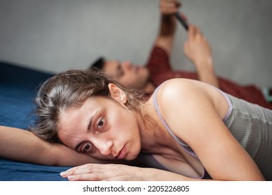 A Young Sad Woman Is Next To Her Partner Who Is Texting On The Phone With Someone, And Not Paying Attention To Her.