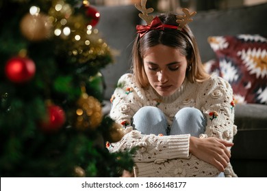 Young sad woman sitting near the Christmas tree. Depressed woman during Christmas holiday at home.