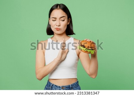 Young sad woman wear white clothes hold eat burger show hand stop gesture say no isolated on plain pastel light green background. Proper nutrition healthy lifestyle fast food unhealthy choice concept