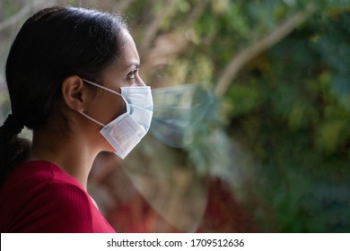 Young sad woman wearing a face mask looking through the window with her reflection on the glass, self isolated at home. Coronavirus and Quarentine concept