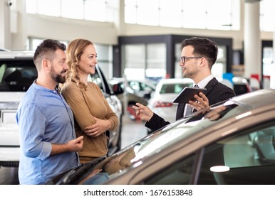 Young Sales Agent Helping Adult Couple To Choose A New Car In Modern Car Showroom.