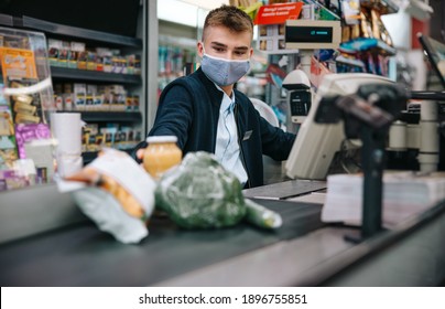 Young Sales Clerk Wearing Face Mask Sitting By Cash Register In Supermarket And Serving Shoppers. Male Cashier Scanning Grocery Products At Checkout.