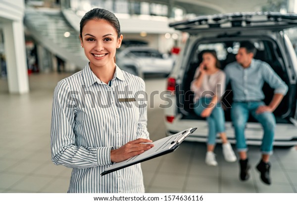 Young saleswoman consultant at a car dealership. In\
the background, the couple is sitting in the open trunk of their\
new car.
