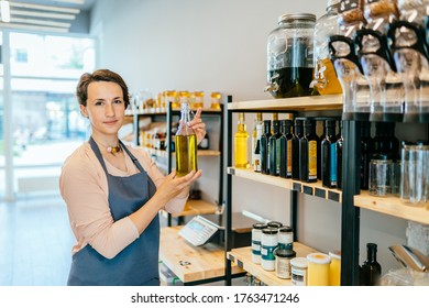 Young saleswoman selling superfoods in zero waste shop. Female owner in apron holds olive oil in glass bottle, stand in grocery store. No plastic conscious minimalism vegan lifestyle concept. - Powered by Shutterstock