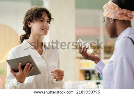 Young saleswoman with tablet looking at female customer with plastic bottle containing liquid self care product and consulting her
