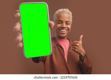 Young satisfied African American woman holds out smartphone with green screen to show off installation of trendy phone application stands on isolated brown background. Mock up, template, copy space