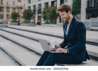 Young Satisfied Businessman In Stylish Dark Blue Suit Sitting On Stairs Near Office Center Outside With Laptop And Coffee To Go In Sunny Summer Day, Working Remotely Online While Having Lunch Break