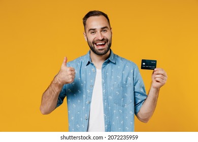 Young Satisfied Cool Smiling Happy Man 20s Wearing Blue Shirt White T-shirt Hold In Hand Credit Bank Card Show Thumb Up Isolated On Plain Yellow Background Studio Portrait. People Lifestyle Concept