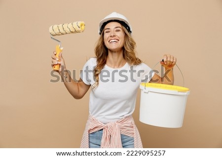 Young satisfied employee laborer handyman woman in white t-shirt helmet use paint roller hold bucket isolated on plain beige background Instruments accessories for renovation room Repair home concept