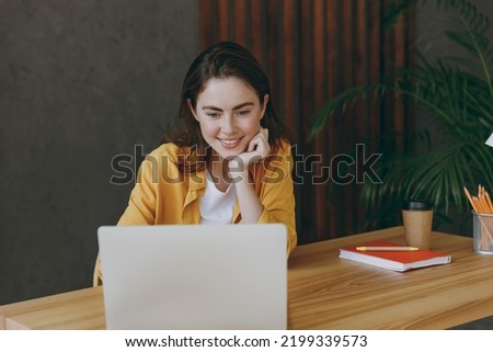 Young satisfied fun happy successful employee business woman 20s she wearing casual yellow shirt hold use laptop pc computer sit work at wooden office desk with pc laptop. Achievement career concept