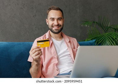 Young Satisfied Fun Man In Casual Clothes Using Work On Laptop Pc Computer Credit Bank Card Shopping Online Order Delivery Booking Tour Sitting On Blue Sofa At Home Flat Indoors Rest Relax On Weekends